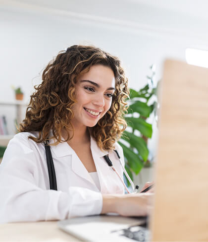 A female doctor wearing a white coat and stethoscope is smiling while working on a laptop in a bright office.