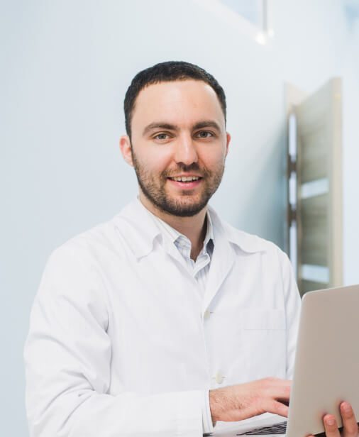 A man in a white lab coat stands indoors, holding and typing on a laptop, looking at the camera and smiling.
