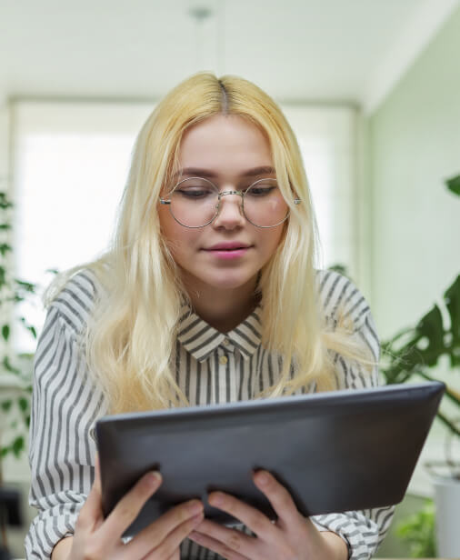 A woman with long blonde hair and glasses, wearing a striped shirt, looks at a tablet indoors with plants in the background.
