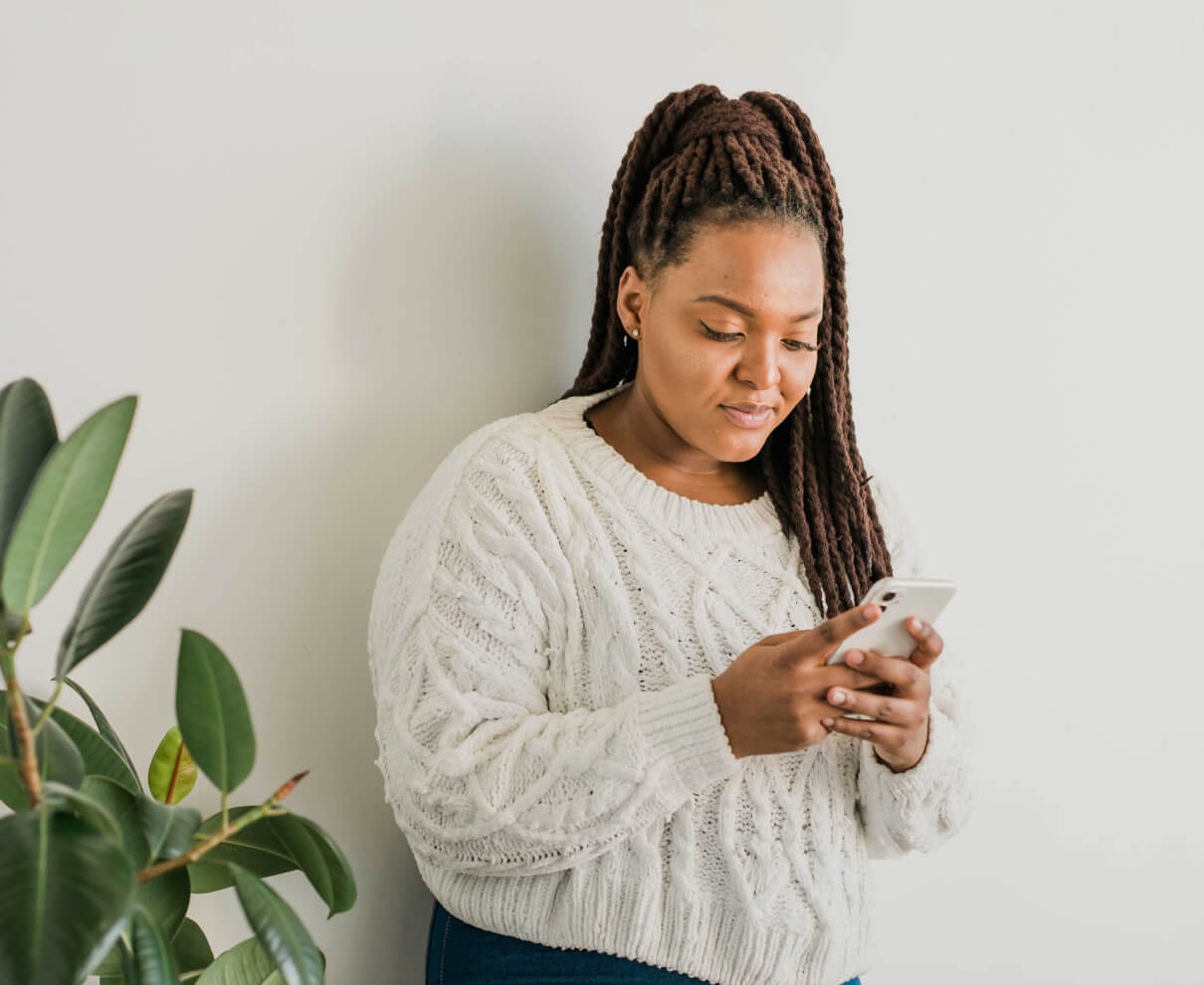 A woman wearing a white sweater stands indoors, looking at and using a smartphone. A green potted plant is visible on her left side.