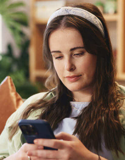 A woman with long brown hair wearing a headband looks at her smartphone while sitting indoors.