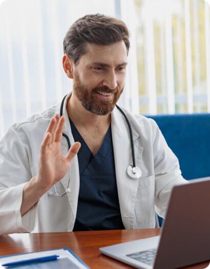 A male doctor wearing a white coat and stethoscope sits at a desk, smiling and waving at a laptop during a virtual consultation.