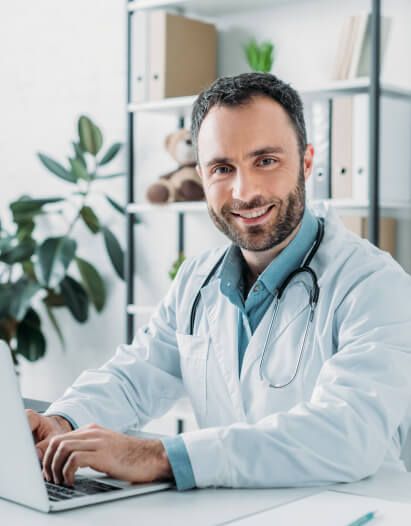A male doctor in a white coat with a stethoscope around his neck is sitting at a desk, smiling, and typing on a laptop in a modern office.