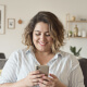 A person with curly hair in a light shirt is smiling while looking at a smartphone in a cozy, well-lit living room.