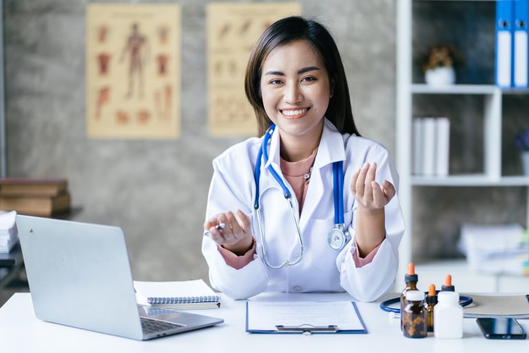 A female doctor sits at a desk with a laptop, clipboard, and medical supplies, smiling and gesturing with her hands in a medical office—showcasing the benefits of integrated healthcare systems for better patient outcomes.