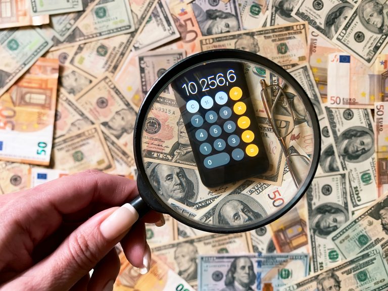 A hand holds a magnifying glass over a smartphone calculator displaying "10,256.6" on top of scattered US dollar bills, highlighting financial details in this sample post.
