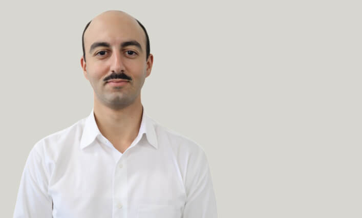 A man with a mustache, wearing a white collared shirt, stands against a plain light gray background.